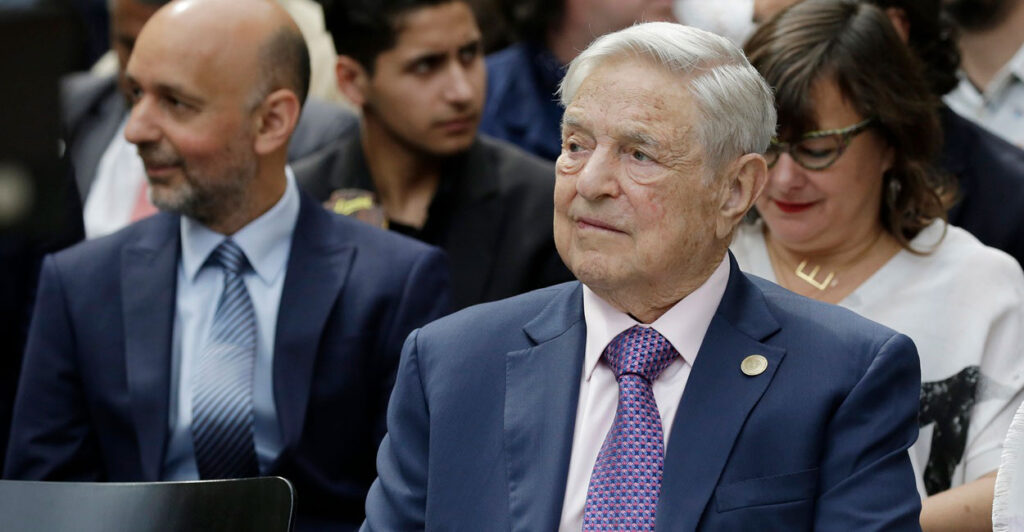 George Soros sits in a chair at an event and looks to ihs left.