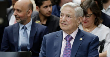 George Soros sits in a chair at an event and looks to ihs left.