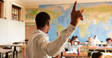 Black teacher in white shirt with back to the camera points to the sky while teaching young students.