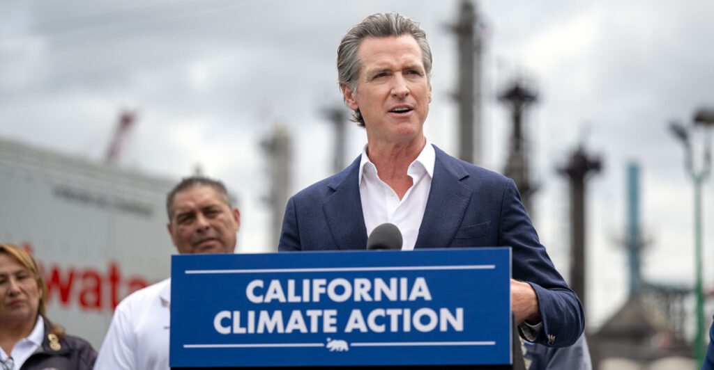 Gavin Newsom, in a blue jacket and white, open-collar shirt, stands at a blue podium with white lettering, reading "California Climate Action."