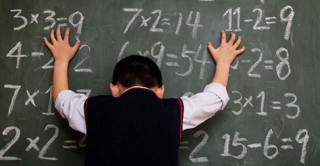 White little boy in a school uniform puts both hands on blackboard featuring simple multiplication tables.