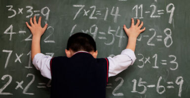White little boy in a school uniform puts both hands on blackboard featuring simple multiplication tables.