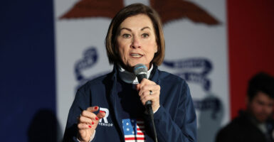 Iowa Gov. Kim Reynolds, in a blue sweater with American flag, speaks into a microphone.