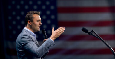 A profile shot Charlie Kirk gesturing with a large American flag in the background.
