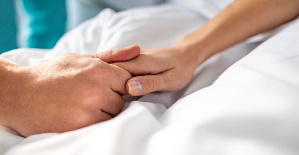 Hand of a man holding the hand of a woman who is in bed with white sheets.