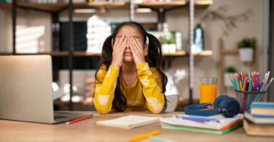Young girl at her school desk with yellow shirt has hands over her eyes.