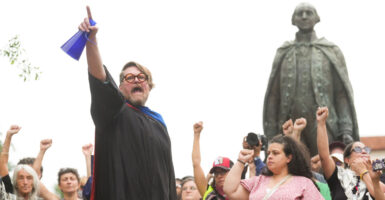 A bearded professor in black robe raises fist surrounded by students and faculty also with raised fist at May Day protest.
