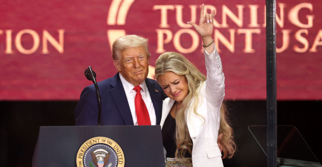 Donald Trump with an arm around Erika Kirk at Charlie Kirk memorial. Erika has her hand in the air and head down.
