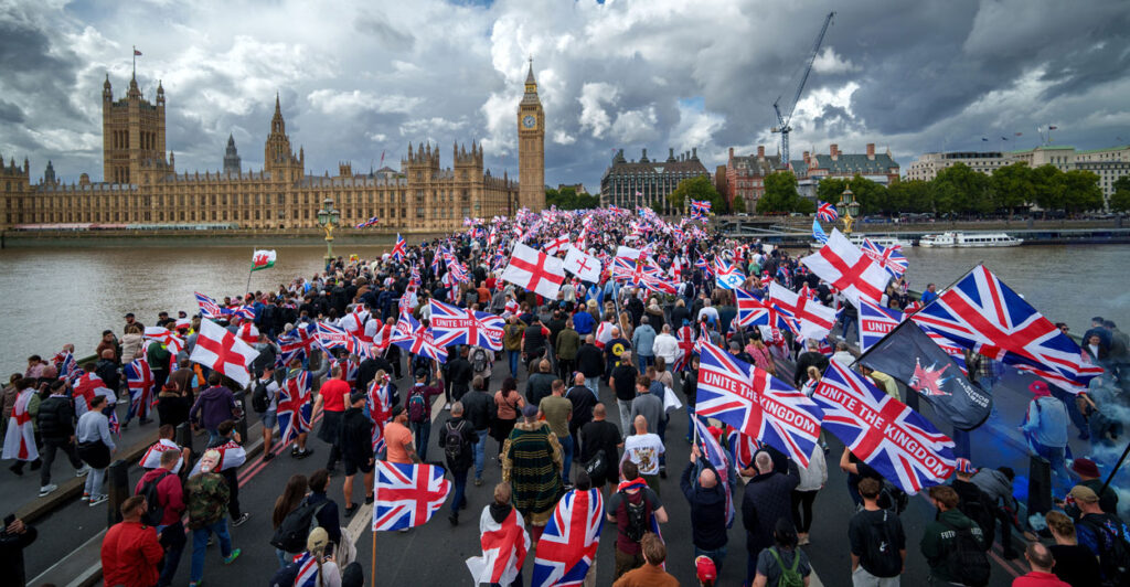 Massive crowd waving Union Jacks and St. George's England flags at protest with Parliament in the background.