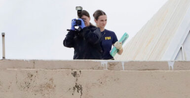 An FBI agent points a camera from the roof of a building, as another agent with documents in her hand stands behind her.