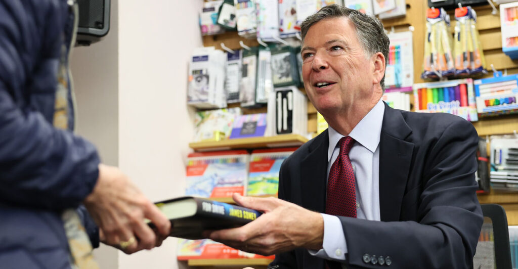 James Comey, seated, hands a copy of his book to someone during a book signing.