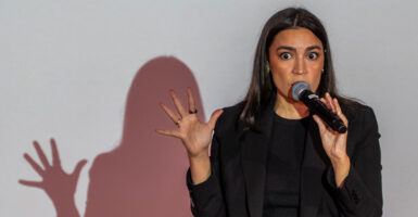 Rep. Alexandria Ocasio-Cortez, D-N.Y., in a spotlight with her shadow on the wall behind her, speaking into a handheld microphone.