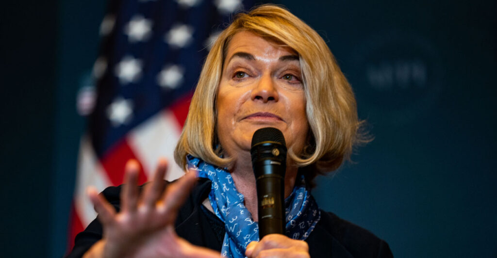 Senator Cynthia Lummis of Wyoming speaks at a political event in front of an American flag back drop, holding a microphone.