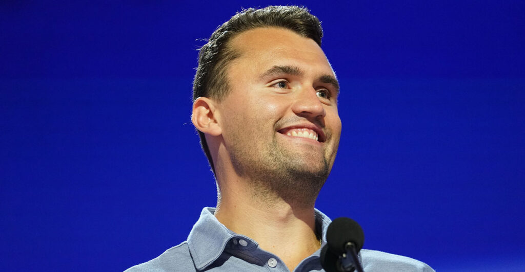 Charlie Kirk smiles in front of a blue background.