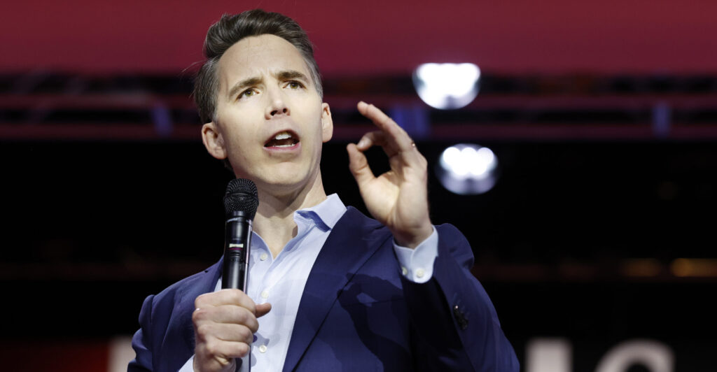 Sen. Josh Hawley in blue jacket and light-blue open shirt gestures as he speaks into a microphone.