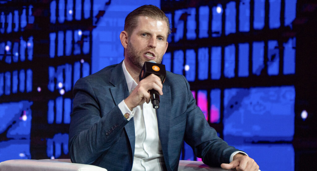 Eric Trump holds a microphone sitting on a white sofa and looking straight to the camera.
