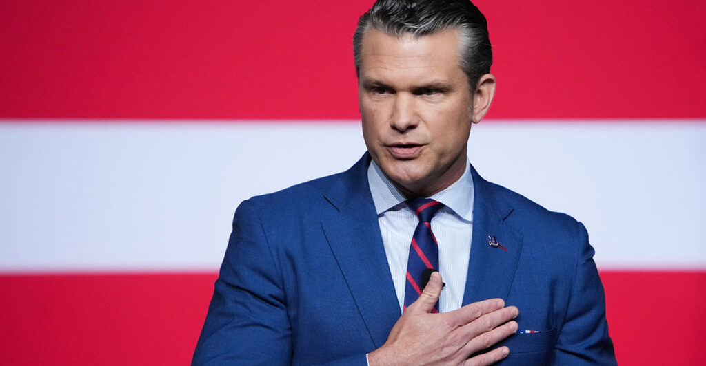 Pete Hegseth in a blue suit puts his hand over his heart.
