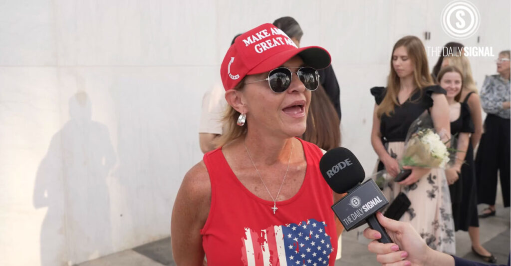 A woman wearing a red Make America Great Again hat a red t-shirt with an American flag on it, speaks to an interviewer at Charlie Kirk's vigil at The Kennedy Center in Washington, D.C.