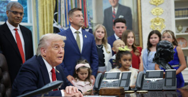 President Donald Trump at his desk in the Oval Office surrounded by childhood cancer survivors and family members.