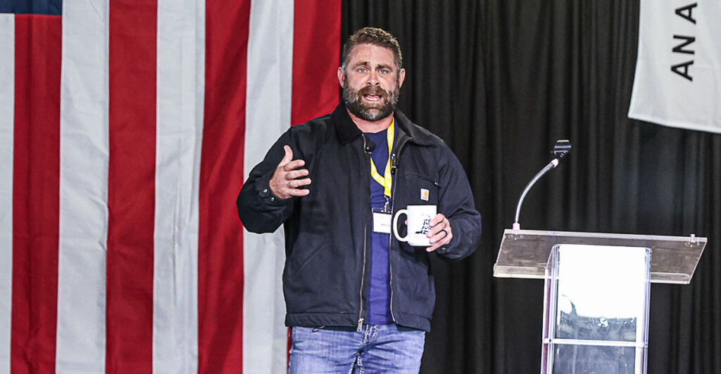 Nick Freitas in a pullover on stage talking to an audience in a warehouse with a large American flag in back