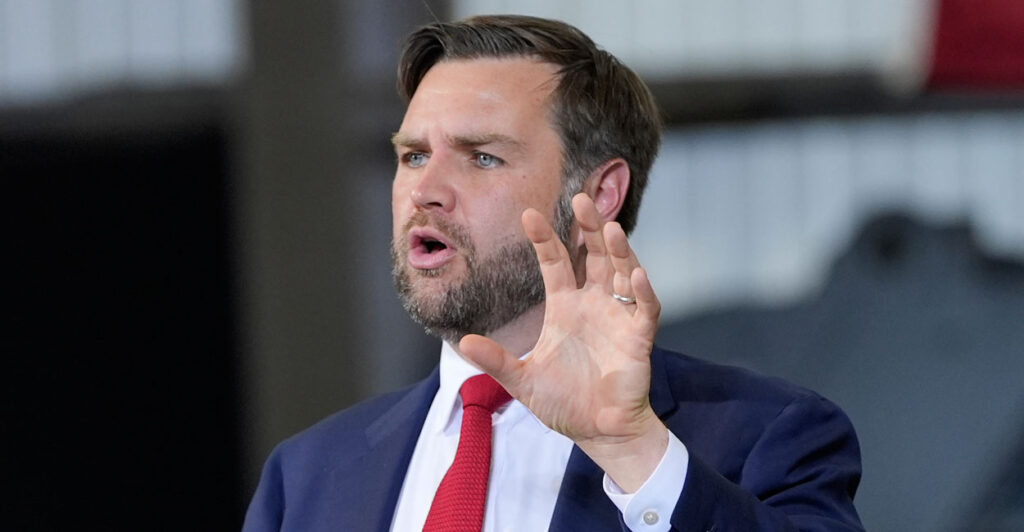 VP JD Vance in a blue suit and red tie gestures with his hand as he speaks to an audience