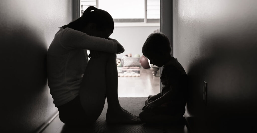 A young woman sitting on the floor of a narrow hallway with her small son with her head to her knees looking to be in despair