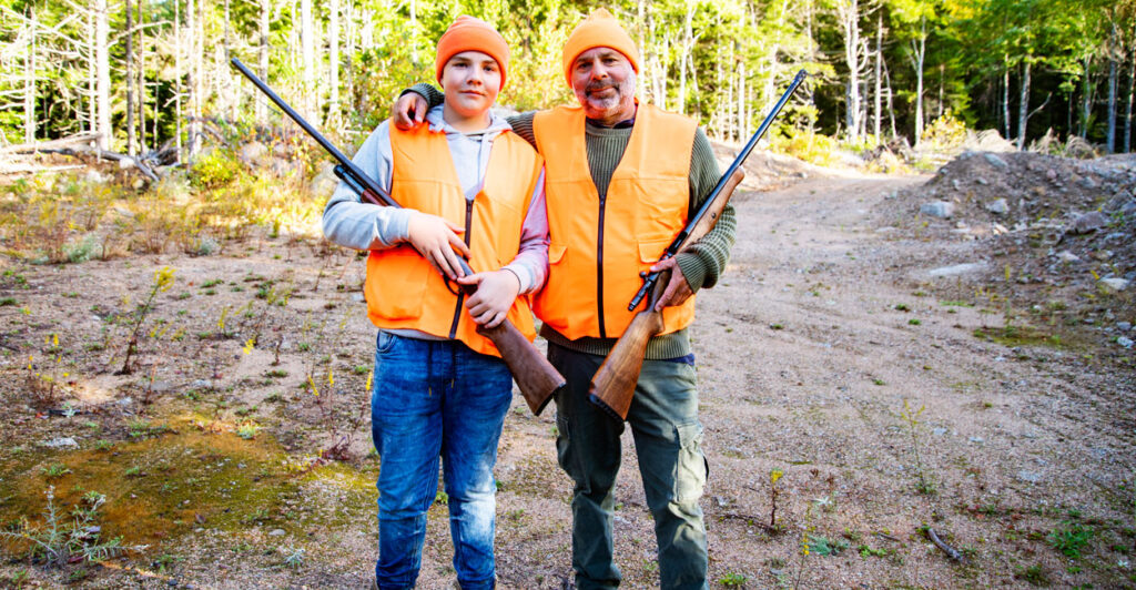 Father and son in orange hunting vests with hunting rifles in rural wooded area.