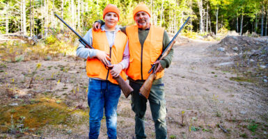 Father and son in orange hunting vests with hunting rifles in rural wooded area.