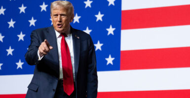 Donald Trump stands in front of an American flag as he points with his right hand.