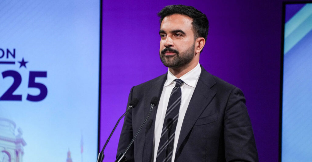 Zohran Mamdani stands behind a podium with a microphone in front of him and looks to his right.