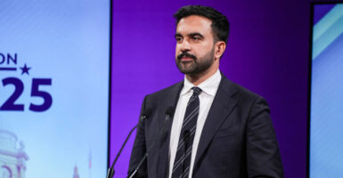Zohran Mamdani stands behind a podium with a microphone in front of him and looks to his right.