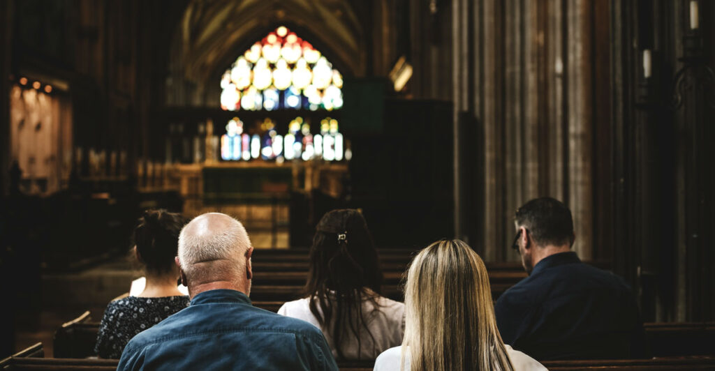 A view of a stained-glass behind the podium at a church, with parishioners in the pews, as seen from behind.