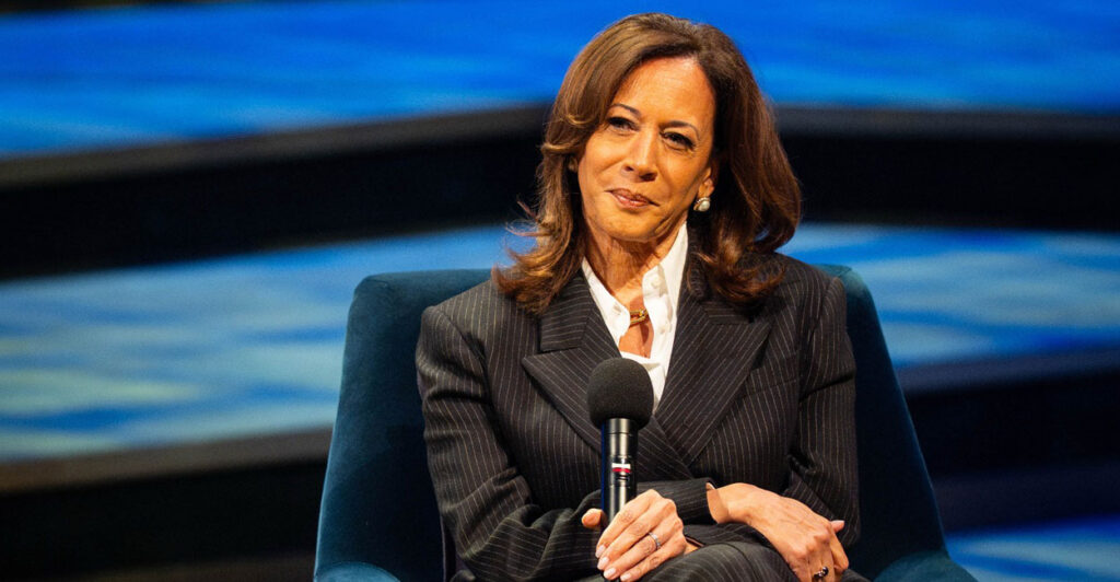 Kamala Harris sits on a chair with her hands folded together and a microphone in her right hand as she listens to a question.