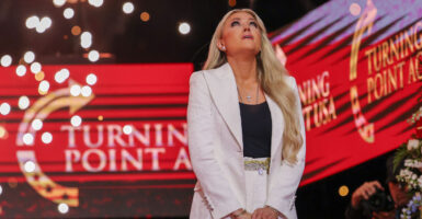Erika Kirk, in white suit and black shirt, looks heavenward, with red Turning Point USA backdrop and sparkles around her.