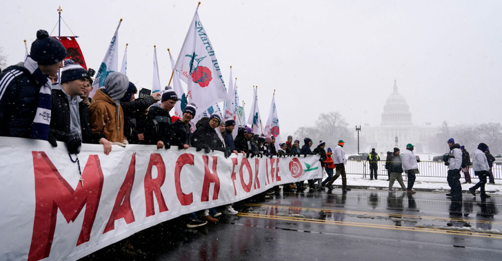 Young people carry the March for Life banner, with the U.S. Capitol in the background on a cold, snowy day.