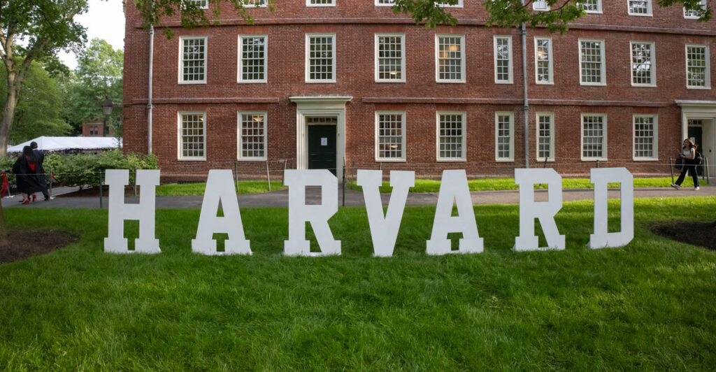 White letters spelling out "Harvard" on grass in front of a brick building.
