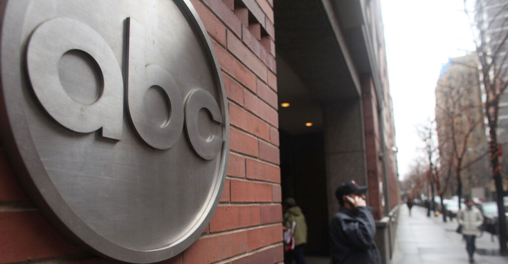 The ABC logo in silver on a brick building, as man stands with cellphone on sidewalk.