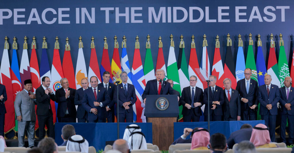 President Trump backed by international leaders and a row of international flags and backdrop reading "Peace in the Middle East," speaks.