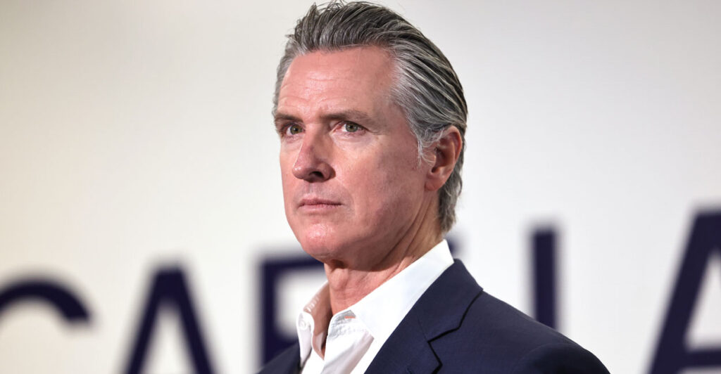 Gavin Newsom in dark jacket and open collar white shirt looking stern.