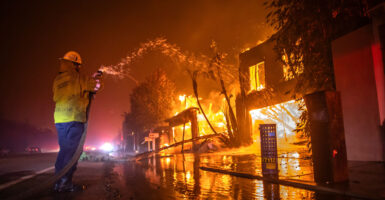 A lone firefighter pours water on a row of buildings totally engulfed in flames at night.