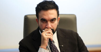 Zohran Mamdani staring into the camera lens, right hand at his mouth.