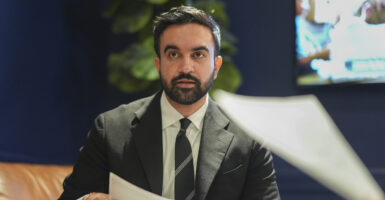 Zohran Mamdani in a dark suit and tie holds a paper in his hand.