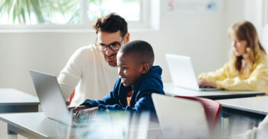 A teacher in white sweater assists a young black student working on a computer, as white girl works at her computer in background.