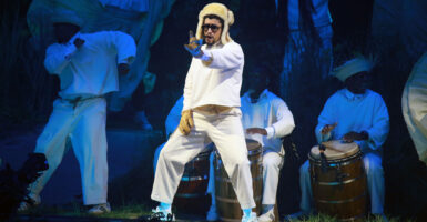 Bad Bunny, in a white outfit with fur hat, performs. Behind him, musicians with bongos.