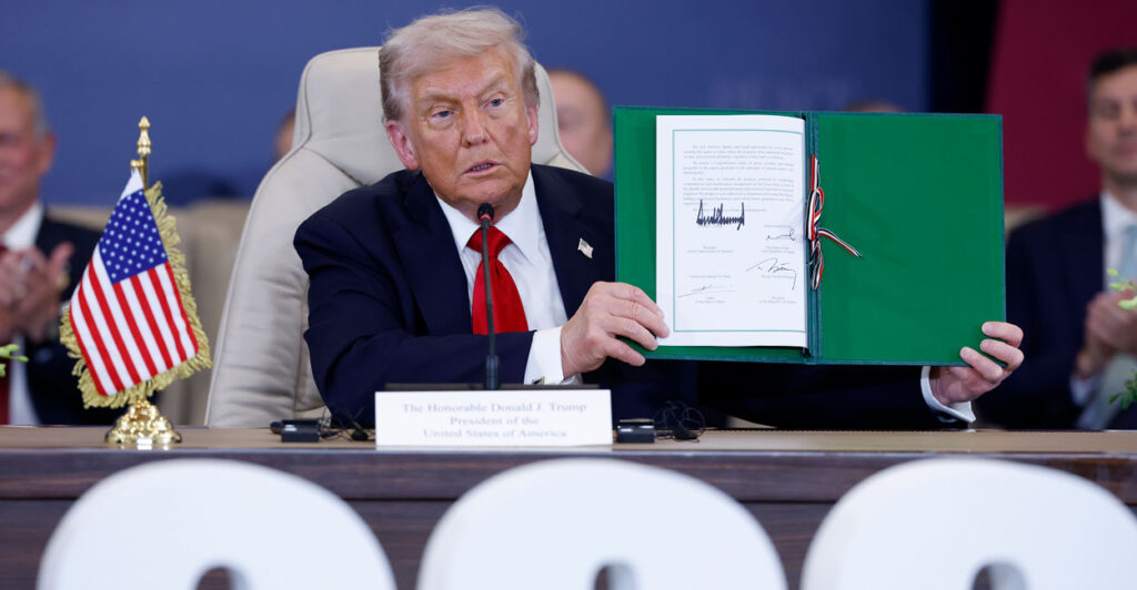 President Trump holds up the signed Gaza peace agreement, while sitting down.
