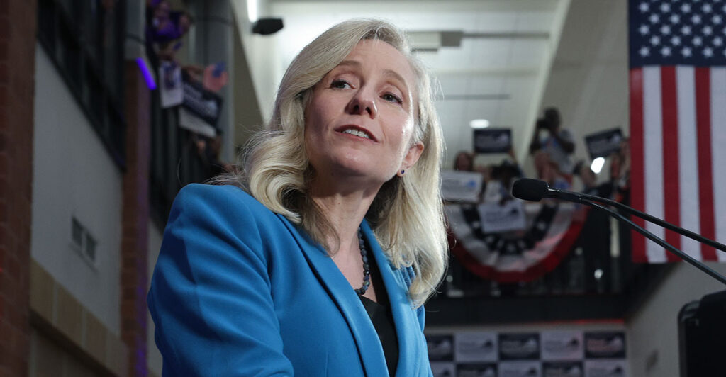 Abigail Spanberger wears blue and speaks in front of American flags.