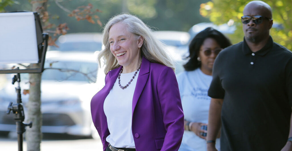 Democrat Abigail Spanberger smiles while wearing a white blouse with a blue sport coat in front of cars.