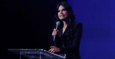 Turning Point USA podcast host Alex Clark on stage speaking into a handheld microphone in a black suit with a dark blue and black background