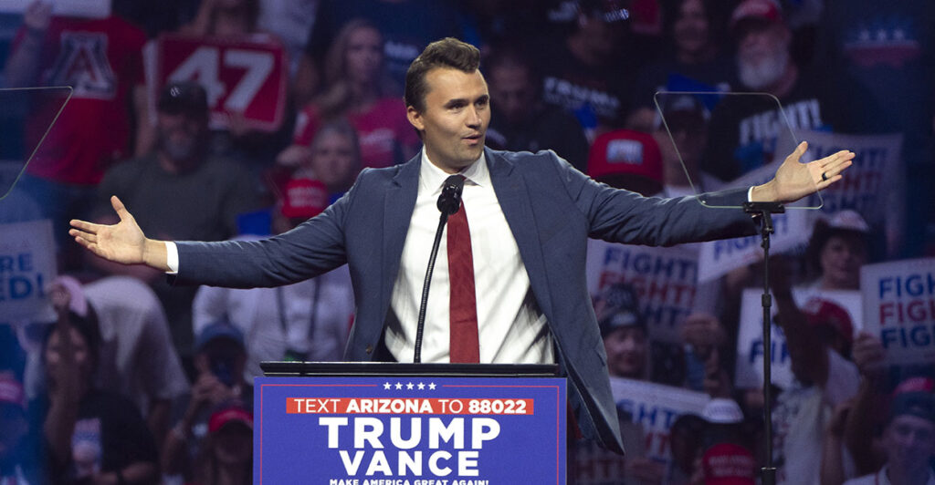 Turning Point USA founder Charlie Kirk in a blue suit with hands outstretched behind a Trump Vance sign.