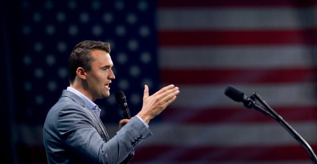 Charlie Kirk gestures as he speaks from a podium with the American flag in the background.